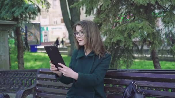 Smiling girl sitting on a bench outdoor, using a tablet outdoor 4k - Felvétel, videó