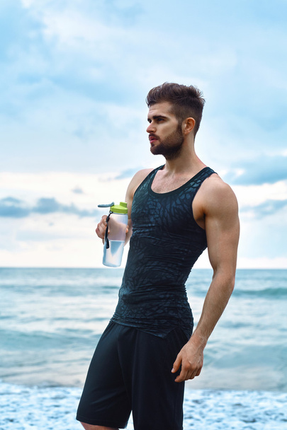 Γυμναστήριο άνθρωπος με μπουκάλι νερό ανάπαυσης μετά από προπόνηση στην παραλία - Φωτογραφία, εικόνα