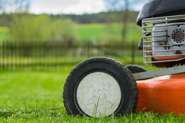 Detail of wheel and piece of motor of lawn mower (grass cutter) on cutted grass with wooden railing, grass field, trees and sky on background. Soft focus / shallow of depth - Foto, Bild