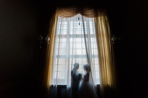 Silhouette of bride and groom embracing holding bouquet against the window with curtains - Photo, Image