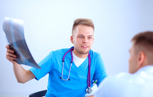 Портрет улыбающегося врача-мужчины с ноутбуком, сидящего за столом в медицинском кабинете
 - Фото, изображение