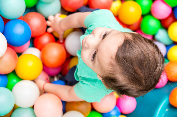 Adorable baby boy wearing turquoise t-shirt playing with colored plastic balls shot from above angle - Fotó, kép