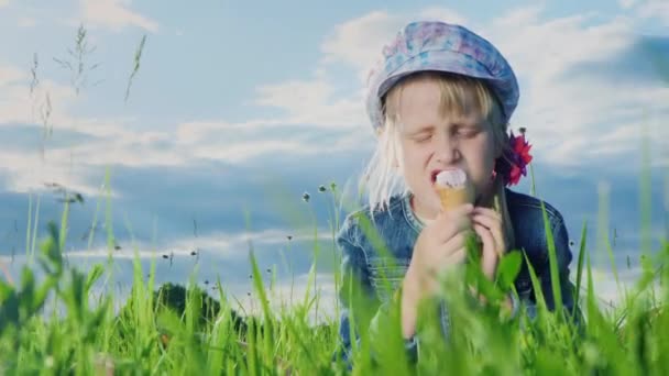 Funny girl eating ice cream outdoors. He sits on the green grass against the sky with beautiful clouds - Кадри, відео