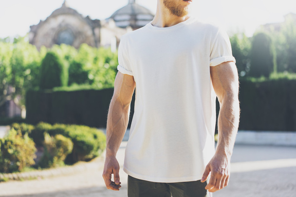 Photo Bearded Muscular Man Wearing White Blank t-shirt. in summer time. Green City Garden Background at sunset. Horizontal Mockup - Foto, afbeelding