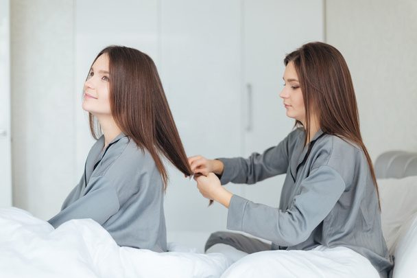 Woman sitting on bed and brushing her sistes twin hair  - Foto, afbeelding