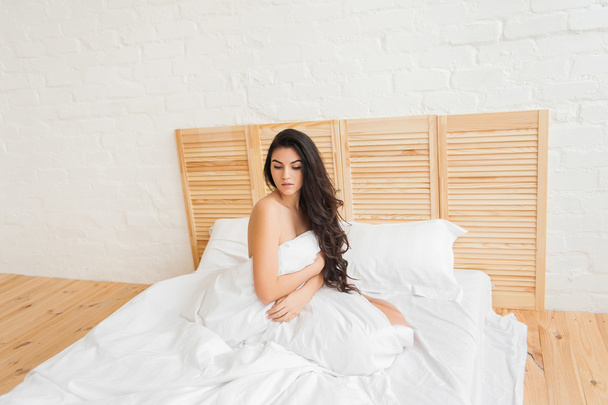 Beautiful brunette sitting on bed with white duvet, looking down - Фото, изображение