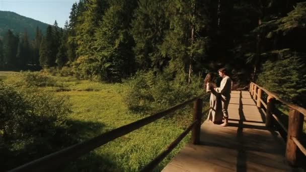 Happy beautiful couple in traditional ukrainian clothes softly kissing and having fun on the wooden bridge in Carpathian mountains - Felvétel, videó