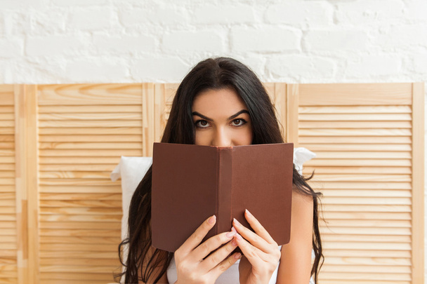 Beautiful young brunette woman hiding herself behind book - Fotó, kép