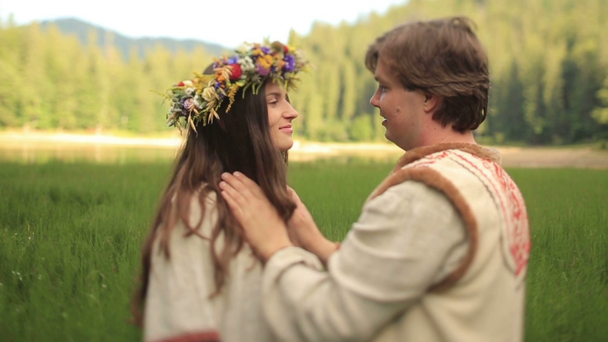 Beautiful couple in love in traditional ukrainian clothes kissing  at the green field. Handsome young man puts on womans head wonderful flower wreath - Materiał filmowy, wideo