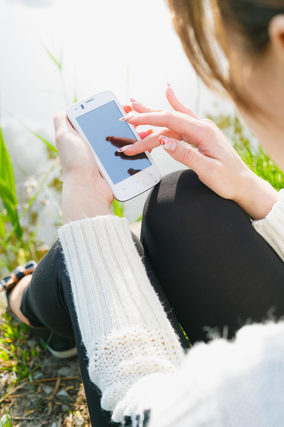 Piękna młoda kobieta za pomocą telefonu komórkowego, siedząc na trawie. Lato zdjęcie. Przybrzeżnych widok. - Zdjęcie, obraz