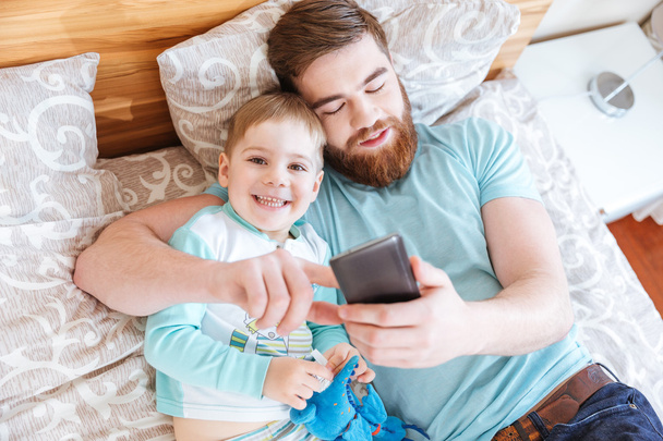 Dad and son using cell phone on bed together - Photo, Image