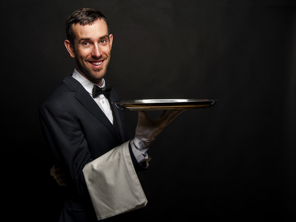 Σερβιτόρος σε μαύρο κοστούμι κρατώντας δίσκο πάνω από το μαύρο φόντο. - Φωτογραφία, εικόνα