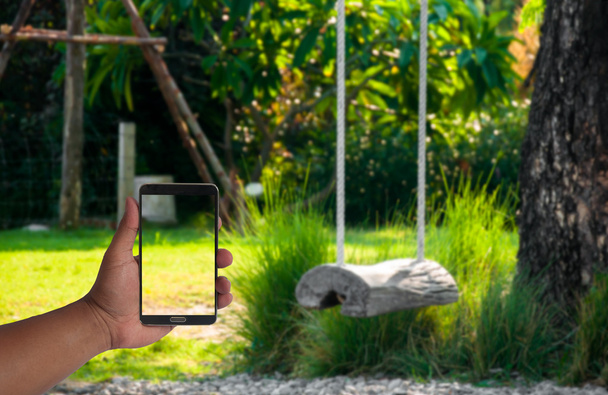 La mano del hombre sostiene el teléfono móvil sobre el oscilación borrosa del registro que cuelga debajo del árbol en el jardín
 - Foto, imagen