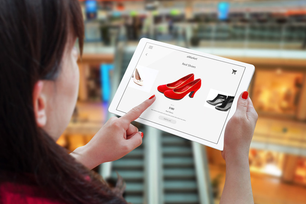 Frau Online-Shopping mit Tablet. Halterung und wählen Sie rote Schuhe. Einkaufszentrum im Hintergrund. - Foto, Bild