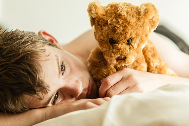 Teini-ikäinen poika käpertyy sänkyyn ruskean nallekarhun kanssa
 - Valokuva, kuva