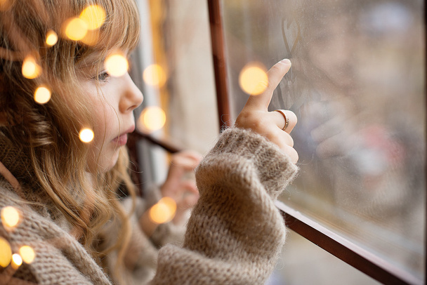 littke hermosa chica con el pelo largo está sentado cerca de la ventana con la decoración de Navidad
 - Foto, Imagen
