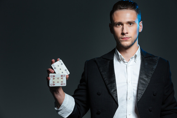 Serious young man magician standing and holding two playing cards  - Fotografie, Obrázek