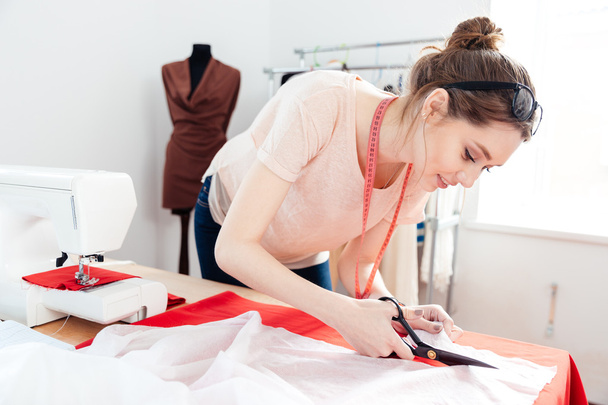 Focused woman fashion designer cutting white fabric in studio - 写真・画像