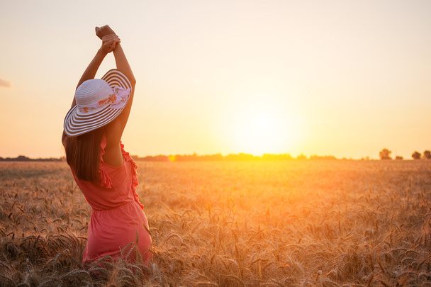 Beautiful young woman with brown hear wearing rose dress and hat enjoying outdoors looking to the sun on perfect wheat field on sunset - Photo, Image