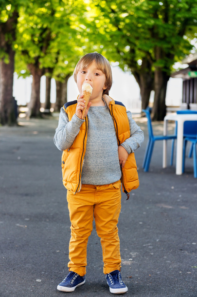 Retrato vertical al aire libre de adorable niño de 4-5 años, con jersey azul, pantalones amarillos y chaleco, comiendo helado en el parque
 - Foto, Imagen