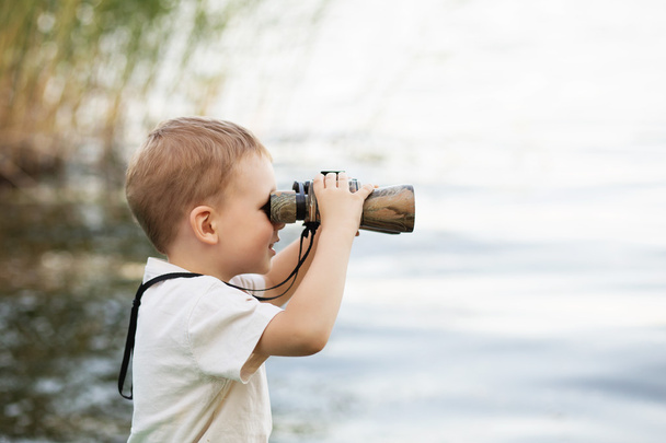 Kleine jongen op zoek door middel van verrekijkers op de rivier bank - Foto, afbeelding
