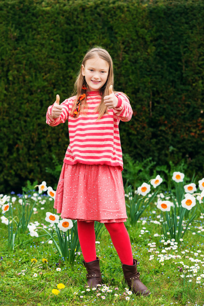 Linda niña de 7-8 años jugando en el parque de primavera entre narcisos flores, pulgares hacia arriba, con jersey de rayas, falda roja y medias
 - Foto, Imagen