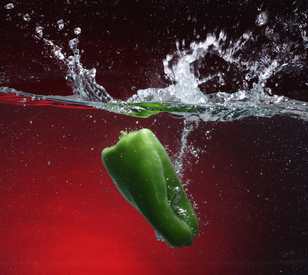 Green pepper falling into water. Red background - Photo, Image