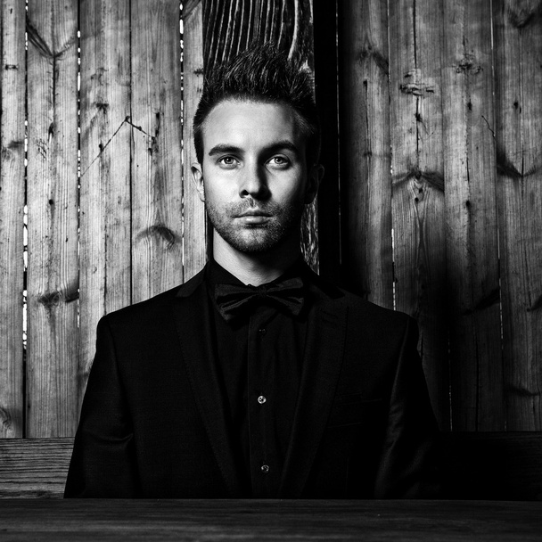 Portrait of young beautiful fashionable man against wooden wall In black suit & bow tie. Black-white fashion photo against wooden background. - Fotoğraf, Görsel