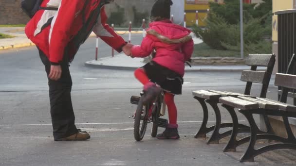 Tatínek pomáhá holčičku jezdit kolo tlačí její běh města ulici večer asfaltové silnici Kid sedí otec muž v červené bundě s batohem - Záběry, video