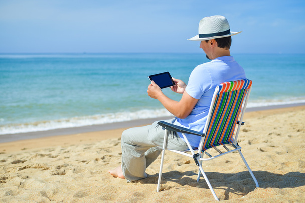 Man using tablet relaxing on ocean beach, blue sky outdoors background - Фото, изображение