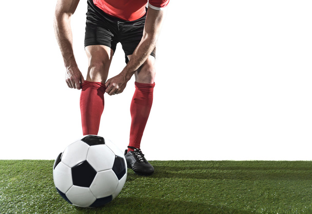 jugador de fútbol con pelota con zapatos negros ajustando su calcetín rojo de pie sobre hierba verde aislado
 - Foto, imagen