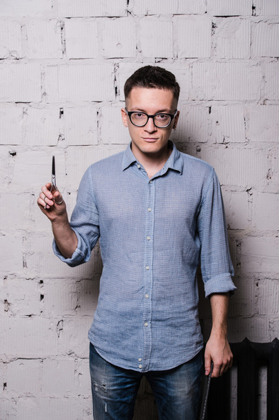 Portrait of young male hairdresser in glasses, posing with scissors, on gray brick wall background, front view, vertical image - Φωτογραφία, εικόνα