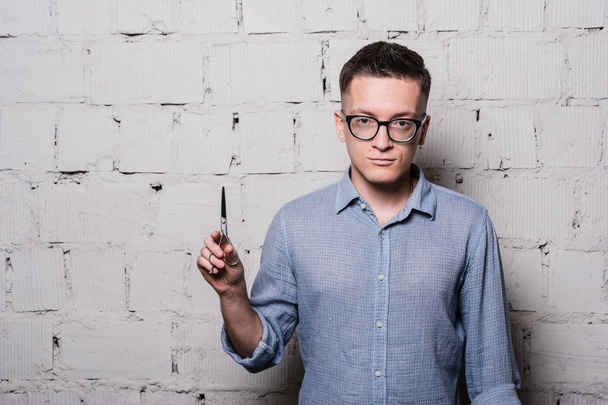 Portrait of young male hairdresser in glasses, posing with scissors, on gray brick wall background, front view, horizontal image - Photo, Image