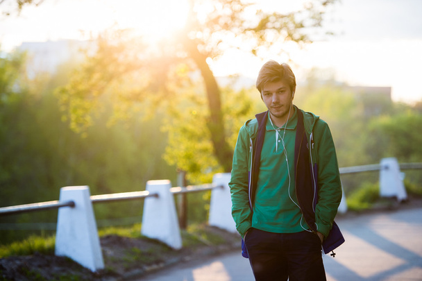 Young man walking around city listening to music with ear headphones looks at the camera in backlight and something ponders. Close-up shot. Sunset. Autumn weather. - Photo, Image