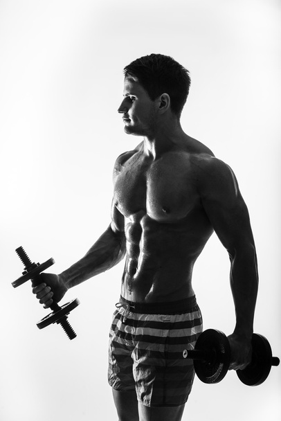 Joven y en forma modelo masculino posando sus músculos bombeando con pesas en un gimnasio mirando a la izquierda aislado sobre fondo blanco
. - Foto, imagen