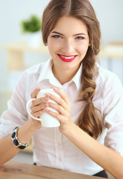 Young businesswoman sitting on the desk with cup in office - Foto, Imagen