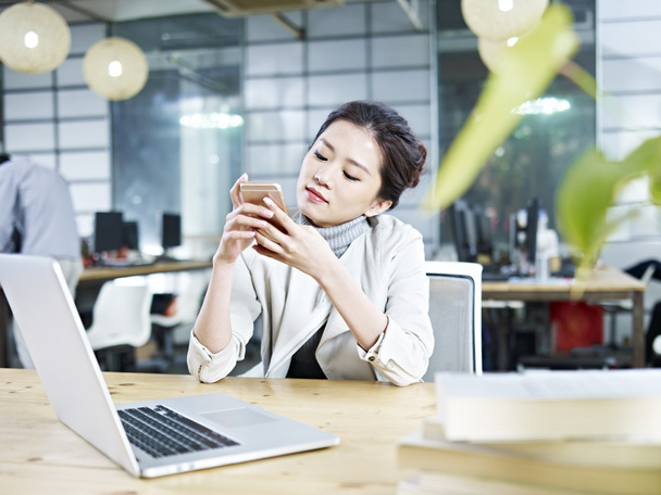 молодая азиатская деловая женщина играет с сотовым телефоном в офисе
 - Фото, изображение