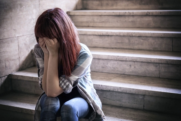 Chica deprimida en crisis adolescente sentada en los escalones de un sótano, cubre su cara con sus manos. Una luz natural penetra desde arriba
. - Foto, imagen