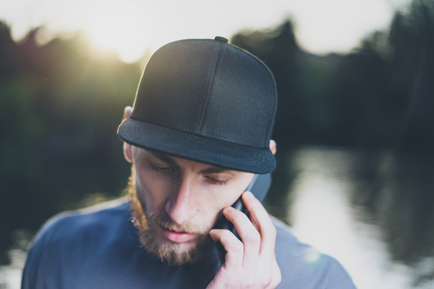 Photo Bearded Young Man Wearing Black Blank Cap Using Modern Smartphone. Green City Park Lake Background and Sunset effect. Front view. Horizontal Mockup - Фото, изображение