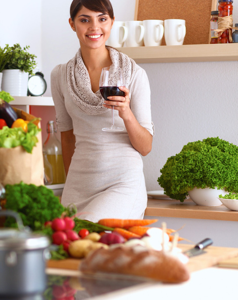 Jeune femme coupant des légumes dans la cuisine, tenant un verre de vin - Photo, image