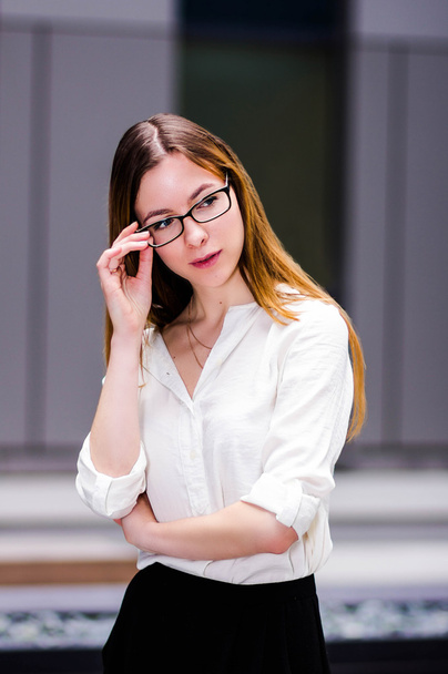 красивая молодая сексуальная стройная и строгая девушка сидит держит очки на фоне бизнес-здания. портретный фон
 - Фото, изображение