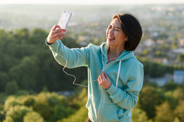 Sensual young japanese woman taking selfie outdoor in park using her phone and laughing - Zdjęcie, obraz