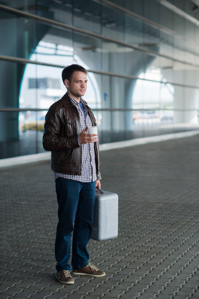 Volledige lengte portret jonge gelukkig lachend knappe reiziger man in 20s verlaten aankomsten luchthavens lounge terminal gebouw na het verzamelen van zijn bagage met een kop verse koffie - Foto, afbeelding
