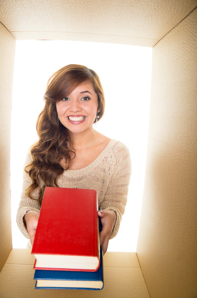 Nice and smiling girl holding two books, red and blue. Inside a cardboard box and white background - 写真・画像