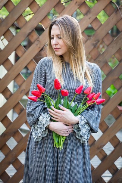 Beautiful young blonde woman with tulips bouquet on against the background of a wooden fence - Foto, Imagen