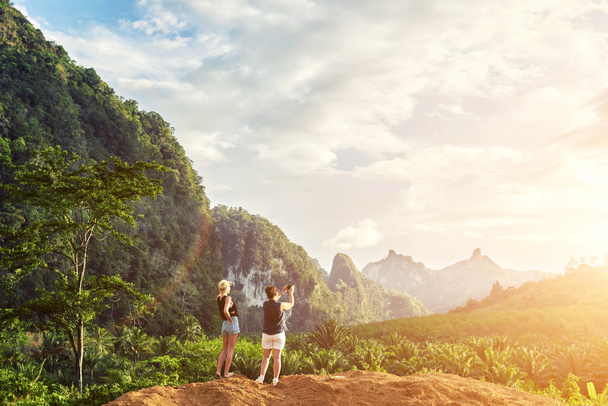 Duas mulheres estão fazendo foto no telefone celular, enquanto está de pé contra a paisagem da selva e céu nublado fundo com espaço de cópia
 - Foto, Imagem
