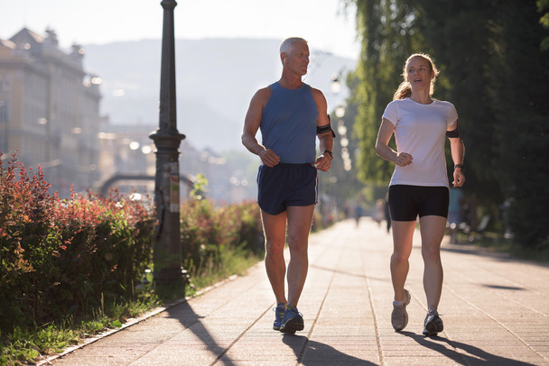 Sağlıklı olgun bir çift sabahın erken saatlerinde arka planda gündoğumuyla birlikte şehirde koşuyor. - Fotoğraf, Görsel