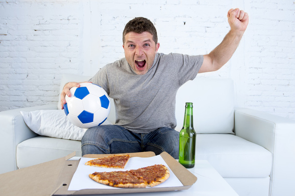 young man holding ball watching football game on tv at home couch with pizza and beer celebrating crazy goal or victory - Photo, Image