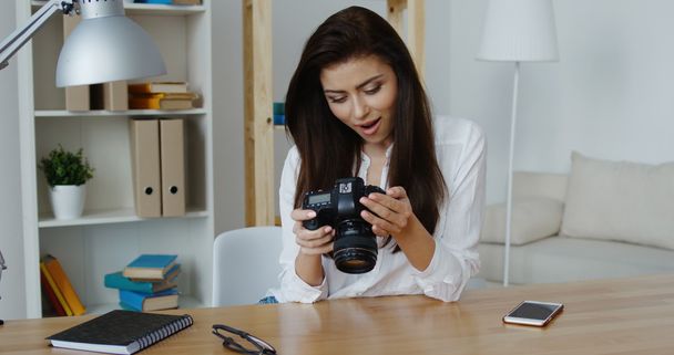 Όμορφη χαμογελαστό πρόγραμμα επεξεργασίας φωτογραφιών μελαχρινή σε λευκό πουκάμισο που βλέπουν μια ψηφιακή φωτογραφική μηχανή στο γραφείο της - Φωτογραφία, εικόνα