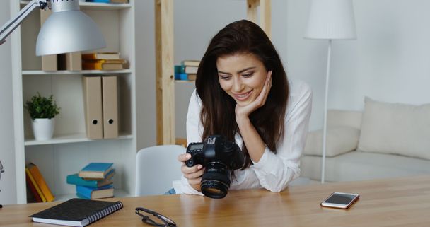Linda morena sorridente editor de fotos em camisa branca olhando para uma câmera digital em seu escritório
 - Foto, Imagem